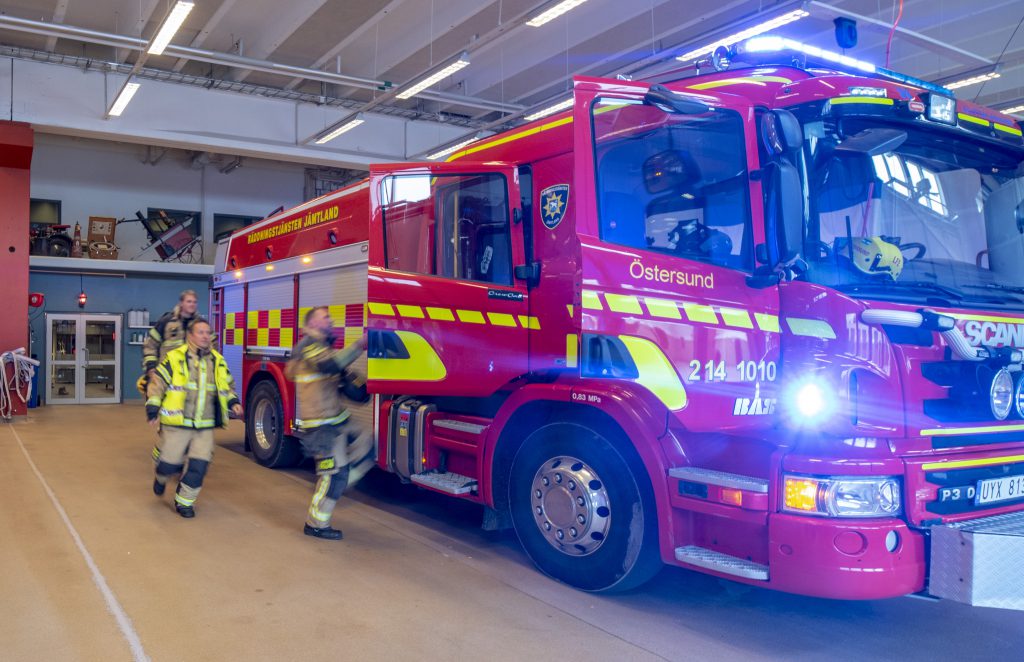 Några brandmän springer mot en röd brandbil i en vagnhall. Brandmännen är suddiga av farten.