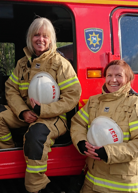 Två glada brandkvinnor i beigea larmställ står framför en bandvagn i rött!