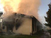 Kameran är riktad mot gaveln på ett flerbostadshus i gulbeige färg. Det ryker från byggnaden och är sotigt från ett litet fönster på nedre våningen.