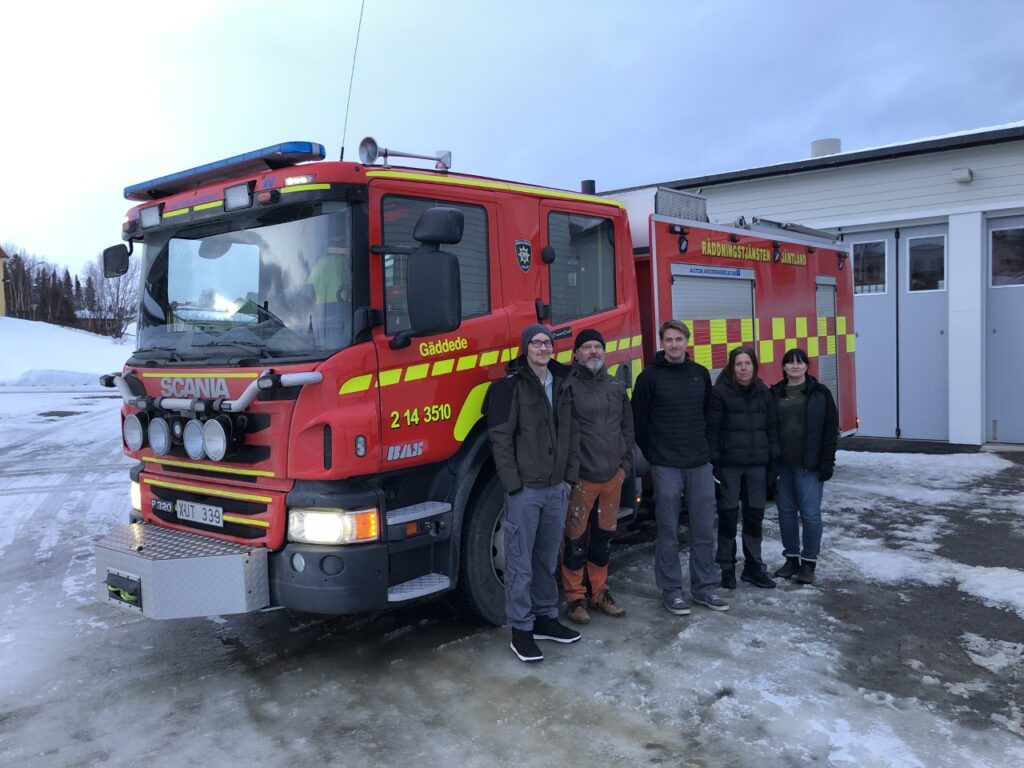 En röd brandbil med fem glada brandmän i civila kläder.